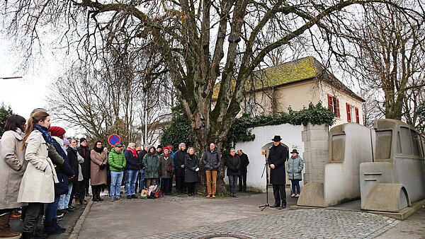 Ein schwarz gekleideter Mann steht rechts vor einem grauen Beton-Bus und hält dort eine Rede. Auf dem gepflasterten Platz vor ihm sind in einem Halbkreis links um ihn herum dutzende Menschen versammelt und hören ihm zu.