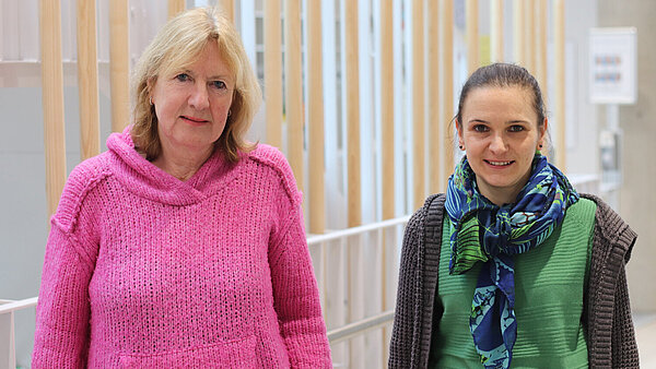 Eine blonde und eine brünette Frau stehen lächelnd in einem hellen Flur.