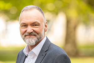 Porträtfoto eines älteren Mannes, im Hintergrund verschwommen der Umriss eines Baumes mit gelben Blättern. Der Mann hat kurze weißgraue Haare und einen Bart. Er trägt ein weißes Hemd mit dunkelgrauem Sakko drüber. Er lächelt mit geschlossenem Mund in die Kamera.