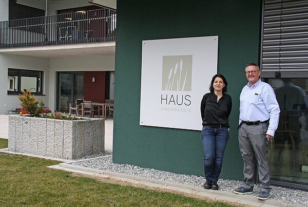 Frau mit dunklen schulterlangen Haaren steht neben älterem Mann mit Hemd und Brille. Beide stehen vor einem petrolfarbenen Gebäude, auf einem Schild steht "Haus Irmengardis". Links neben ihnen sieht man die Terrasse und Balkone des Pflegeheims.