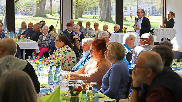 In einem Festsaal, in den die Frühlingssonne scheint, sitzen Seniorinnen und Senioren an festlich gedeckten Tischen; im Hintergrund spricht ein Mann stehend in ein Mikrofon.