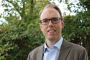 Portraitbild Martin Buchecker, ein junger Mann mit Brille, hellblauem Hemd und beige Sakko. Er lächelt sanft mit geöffnetem Mund.