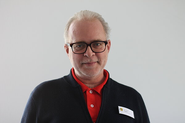 Portrait Bild Thomas Käser. Mann mit grauen Haaren, grauem Bart, schwarzer Brille. Er Trägt ein rotes Polo und eine Strickweste in dunkelblau darüber.