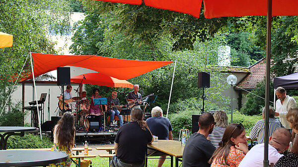 Menschen sitzen auf Bierbänken unter bunten Sonnenschirmen in einem Park, an dessen Ende eine Bühne mit Musikern zu sehen ist. 