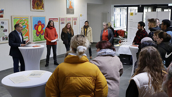 In einem großen hellen Raum hängen Bilder und Infotafeln an den weißen Wänden, das Foto entstand bei der Eröffnung der Ausstellung, im Hintergrund stehen die Redner:innen, im Vordergrund interessierte Zuhörer:innen.