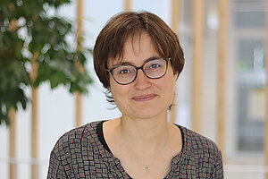 Porträtfoto einer jungen Frau. Sie hat dunkelbraune, kurze glatte Haare. Sie trägt eine runde dunkle Brille, trägt ein graues Langarmshirt und steht vor einem dunkelgrauen Hintergrund. Sie lächelt mit geöffnetem Mund in die Kamera.