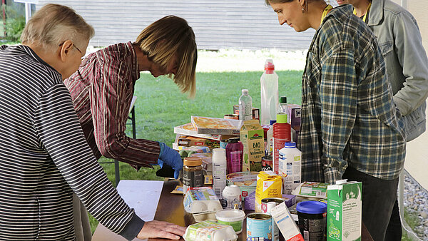 Auf einem Tisch sind mehrere Lebensmittel aufgestellt. Eine Frau mit Handschuhen versucht, passende Münzen aus einem Geldbeutel zu nehmen.