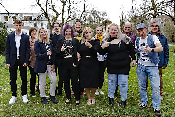 Eine Gruppe von zwölf Frauen und Männern unterschiedlichen Alters steht im Freien auf einer Wiese und schaut Richtung Kamera, alle lächeln, manche machen die Daumen-hoch-Geste.