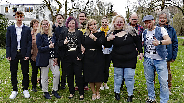 Eine Gruppe von zwölf Frauen und Männern unterschiedlichen Alters steht im Freien auf einer Wiese und schaut Richtung Kamera, alle lächeln, manche machen die Daumen-hoch-Geste.
