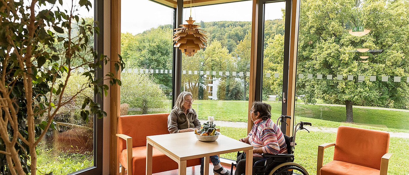 Zwei ältere Damen sitzen sich in einem hellen Aufenthaltsbereich gegenüber, eine der beiden sitzt in einem Rollstuhl. Im Hintergrund öffnet sich der Blick ins Grüne. 