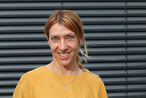 Portrait von einer Frau. Sie trägt eine gelbe Bluse, hat blonde lange Haare und blaue Ausgen.