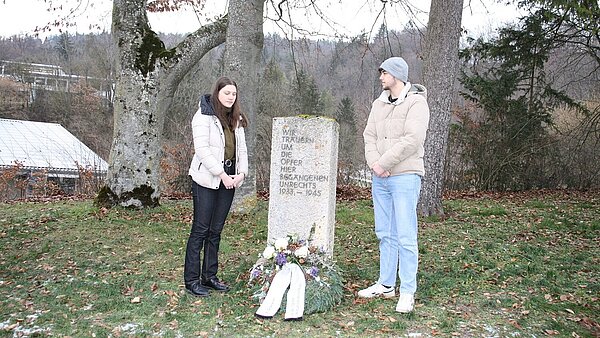 Eine junge Frau und ein junger Mann stehen vor einer Gedenktafel. Vor dieser liegt ein geschmückter Kranz mit Trauerschleife. Beide halten den Blick gesenkt zu Boden. 