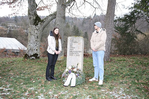 Eine junge Frau und ein junger Mann stehen vor einer Gedenktafel. Vor dieser liegt ein geschmückter Kranz mit Trauerschleife. Beide halten den Blick gesenkt zu Boden. 