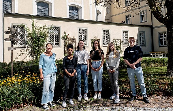 Unter einem schattigen Baum stehen in einer Reihe sechs Personen nebeneinander. Links ist eine Frau zu sehen, rechts neben ihr stehen vier junge Frauen und ganz rechts ein junger Mann. Im Hintergrund befindet sich ein Gebäude mit gelber älterer Fassade.