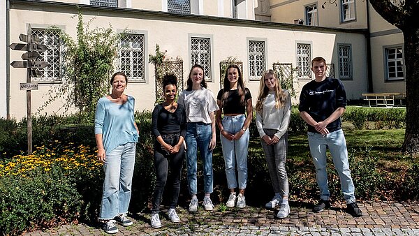 Unter einem schattigen Baum stehen in einer Reihe sechs Personen nebeneinander. Links ist eine Frau zu sehen, rechts neben ihr stehen vier junge Frauen und ganz rechts ein junger Mann. Im Hintergrund befindet sich ein Gebäude mit gelber älterer Fassade.