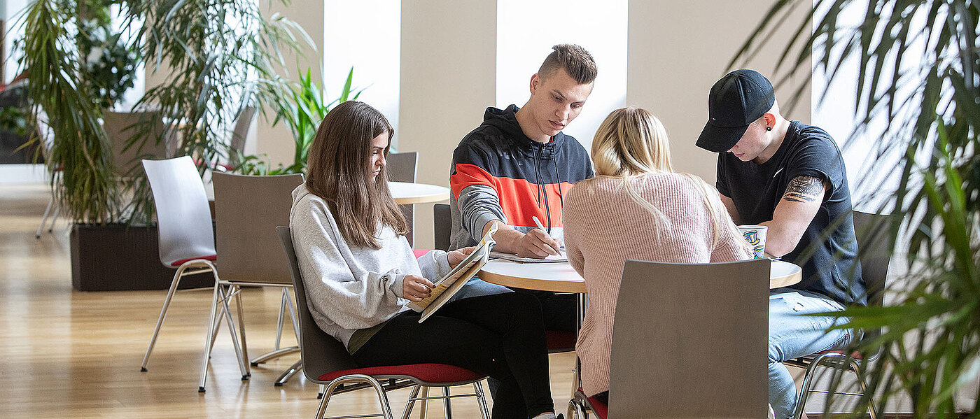 Vier junge Menschen, zwei weiblich, zwei männlich, sitzen gemeinsam bei Tag an einem runden Tisch in einem Flur mit weißen Wänden und hellen großen Fensteröffnungen, sie lesen oder schreiben und sind in ihre Arbeiten vertieft, in dem Flur stehen links von ihnen weitere Tische und Stühle sowie links und rechts von ihnen große grüne Zimmerpflanzen in dunklen rechteckigen Trögen.