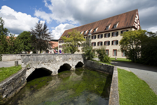 Man sieht ein historisches 3-stöckiges Gebäude, im Vordergrund führt eine steinerne Brücke über einen Fluss.