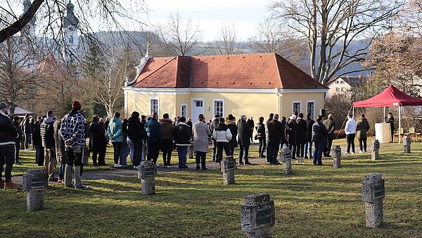 Eine große Gruppe Menschen steht auf einem Friedhof. Im Vordergrund sind einige steinerne Kreuze zu erkennen, im Hintergrund die Türme des Münsters.