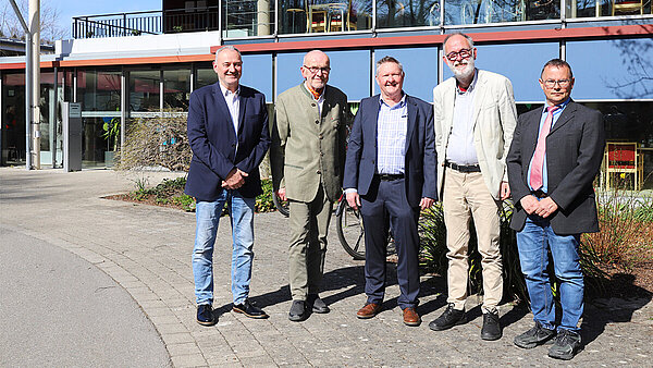 Fünf Männer in Business-Kleidung stehen vor dem Eingang eines Heimgebäudes in der Sonne und lächeln in die Kamera.