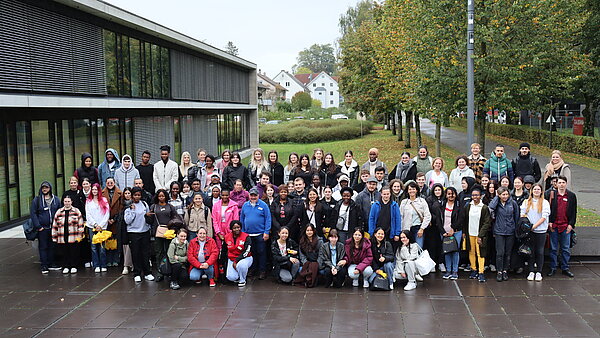 Eine große Gruppe junger Menschen steht in mehreren Reihen hinter- und nebeneinander, im Hintergrund ist ein Gebäude und eine Parkanlage zu sehen. 