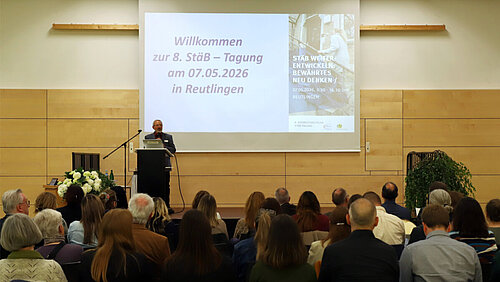 Ein Mann steht in einer Veranstaltungshalle auf einem Podium, vor ihm sitzen zahlreiche Zuhörerinnen und Zuhörer, auf einer Leinwand hinter ihm ist das Tagungsprogramm zu sehen.