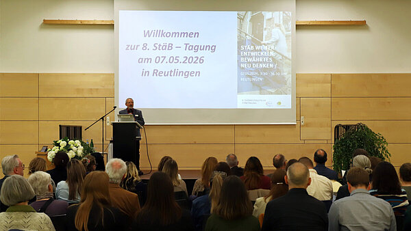 Ein Mann steht in einer Veranstaltungshalle auf einem Podium, vor ihm sitzen zahlreiche Zuhörerinnen und Zuhörer, auf einer Leinwand hinter ihm ist das Tagungsprogramm zu sehen.