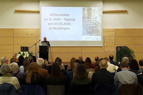Ein Mann steht in einer Veranstaltungshalle auf einem Podium, vor ihm sitzen zahlreiche Zuhörerinnen und Zuhörer, auf einer Leinwand hinter ihm ist das Tagungsprogramm zu sehen.