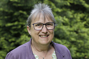 Porträtfoto einer Frau mittleren Alters. Sie hat zusammengebundene grau-weiße Haare mit einem Pony. Sie trägt eine eckige Brille mit dunklem Rahmen, einen violetten Blazer mit Kragen und Bluse darunter sowie eine Kette mit Anhänger um den Hals. Sie lächelt mit geöffnetem Mund in die Kamera. Im Hintergrund sind verschwommen Bäume zu sehen.
