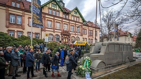 Man sieht eine große Menschengruppe, im Vordergrund ist ein Bus aus grauem Beton, im Hintergrund historische Fachwerkgebäude.  