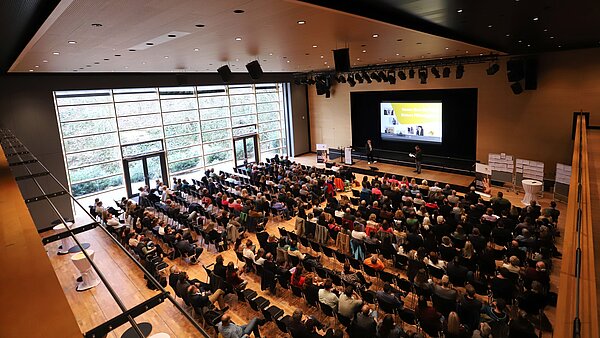 In einem großen Saal sitzen 350 Teilnehmende auf Stühlen, auf der Bühne stehen zwei Personen, die etwas erklären. 