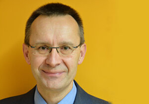 Portraitbild Dr. Andreas Meyer. Er steht vor einem gelben Hintergrund, trägt Brille und lächelt mit geschlossenem Mund.