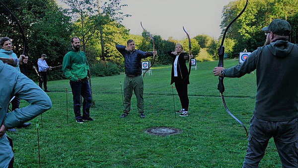 Eine Gruppe Menschen steht auf einer grünen Wiese. Sie halten Pfeil und Bogen in den Händen.