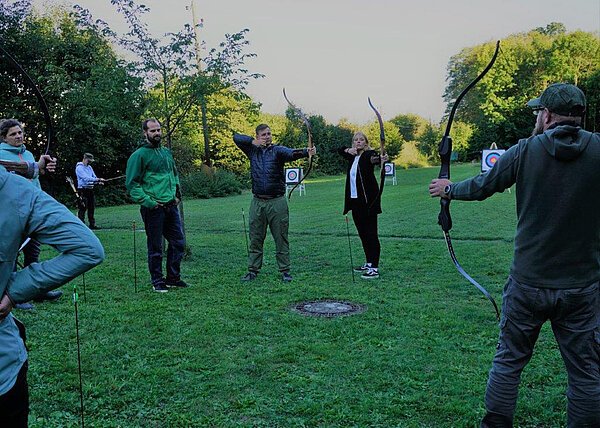 Eine Gruppe Menschen steht auf einer grünen Wiese. Sie halten Pfeil und Bogen in den Händen.