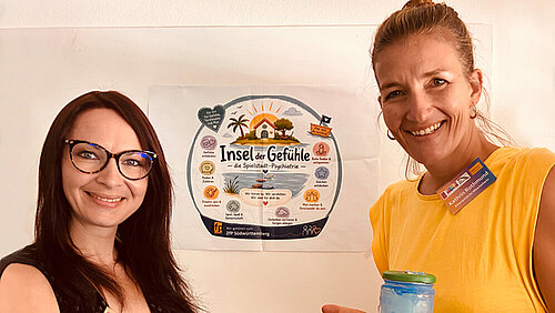 Zwei Frauen stehen vor einem Plakat auf dem "Insel der Gefühle" zu lesen ist.