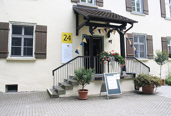 Vorderseite mit Eingangsbereich eines älteren Gebäudes mit dunklen Fensterläden, einer Treppe davor und dunklem Vordach. An der Fassade ist ein gelbes Schild mit der Zahl 24 in schwarz geschrieben. Vor der Treppe stehen Pflanzgefäße und ein Aufstellschild. Am Vordach hängen bunte Stoffwimpel.