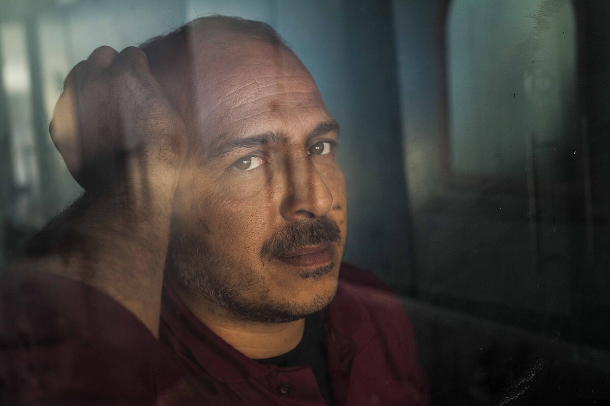 Ein Mann mit Schnurrbart ist hinter einer Scheibe. Er stützt mit der rechten Hand seinen Kopf und schaut nachdenklich.