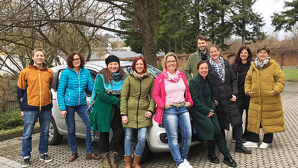 Ein größere Gruppe von Menschen hat sich rund um ein kleines weißes Auto aufgestellt.