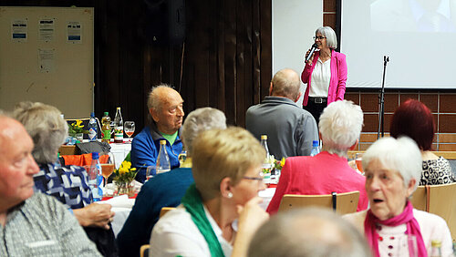 Eine Moderatorin im pinken Blazer spricht mit Mikrofon in der Hand vor an festlich gedeckten Tischen sitzenden Pensionärinnen und Pensionären.