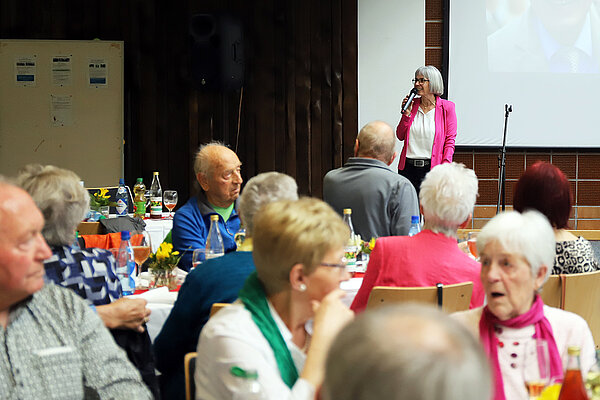 Eine Moderatorin im pinken Blazer spricht mit Mikrofon in der Hand vor an festlich gedeckten Tischen sitzenden Pensionärinnen und Pensionären.