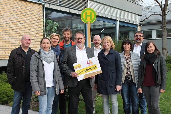 Zehn Personen stehen an einem Busshaltestellen-Schild der Mann mit Brille in der Mitte hält ein Schild in den Händen, auf dem ZfP-Jobticket steht.