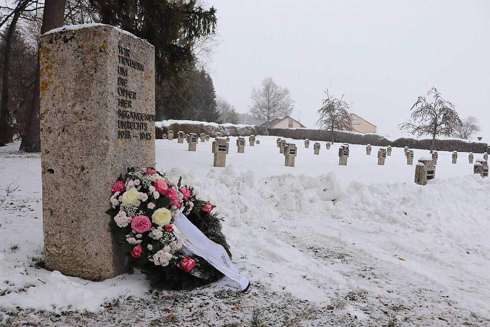 Im Vordergrund sieht man einen rechteckigen Gedenkstein mit einem Blumenkranz davor, im Hintergrund ist ein schneebedecktes Feld mit Grabsteinen zu erkennen.