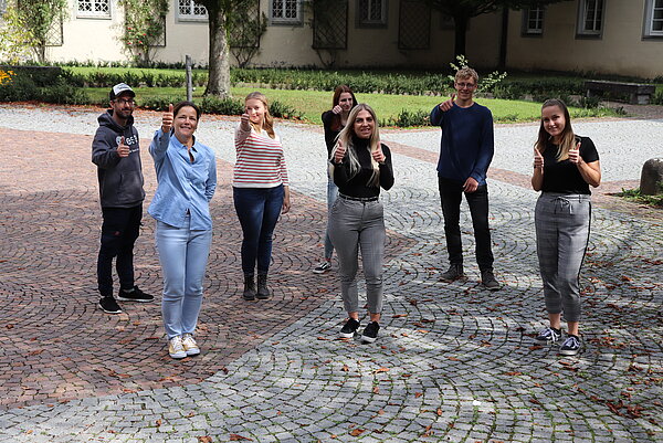 sieben Menschen sind in zwei Reihen versetzt aufgestellt im freien und halten alle den Daumen hoch in die Kamera.