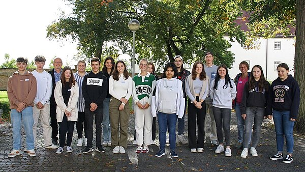 In einer Reihe stehen mehrere junge Menschen mit einem Anleiter und einer Anleiterin nebeneinander und schauen in die Kamera. Die Personen stehen auf einem gepflasterten Platz. Links im Hintergrund ist der blaue Himmel, rechts hinter den Menschen ein großer Laubbaum.