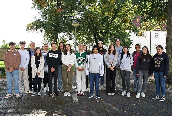 In einer Reihe stehen mehrere junge Menschen mit einem Anleiter und einer Anleiterin nebeneinander und schauen in die Kamera. Die Personen stehen auf einem gepflasterten Platz. Links im Hintergrund ist der blaue Himmel, rechts hinter den Menschen ein großer Laubbaum.