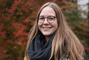 Porträtfoto einer jungen Frau mit blauen Augen und langen braunen glatten Haaren. Sie trägt einen dicken schwarzen Schal und eine runde Brille. Sie lächelt mit geöffnetem Mund in die Kamera. Im Hintergrund sind rot gefärbte Laubbäume zu sehen.