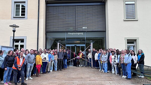 Eine große Gruppe Menschen steht vor einem mehrstöckigen Gebäude, im Hintergrund ist ein Eingang zu erkennen.