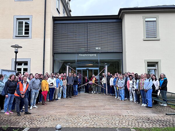 Eine große Gruppe Menschen steht vor einem mehrstöckigen Gebäude, im Hintergrund ist ein Eingang zu erkennen. 