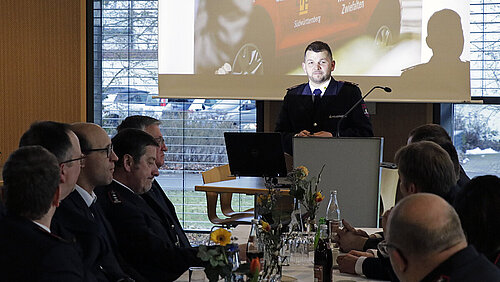 Ein junger Mann in Feuerwehruniform steht vor sitzenden Kamerad:innen an einem Rednerpult, im Hintergrund ist eine PowerPoint-Präsentation zu sehen.