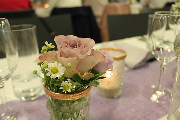 Auf einem festlich gedeckten Tisch stehen Gläser sowie in der Mitte ein Teelicht im Glas und daneben eine Glasvase mit Blumendekoration darin.