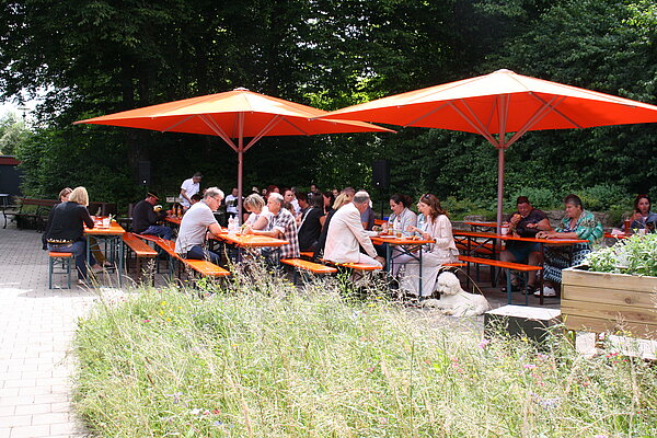 An mehreren Biertischgarnituren sitzen einige Menschen. Sie sitzen unter orange-farbenen Sonnenschirmen. Im Hintergrund sind Bäume zu sehen, im Vordergrund eine Blumenwiese und der gepflasterte Weg zu den Bierbänken.